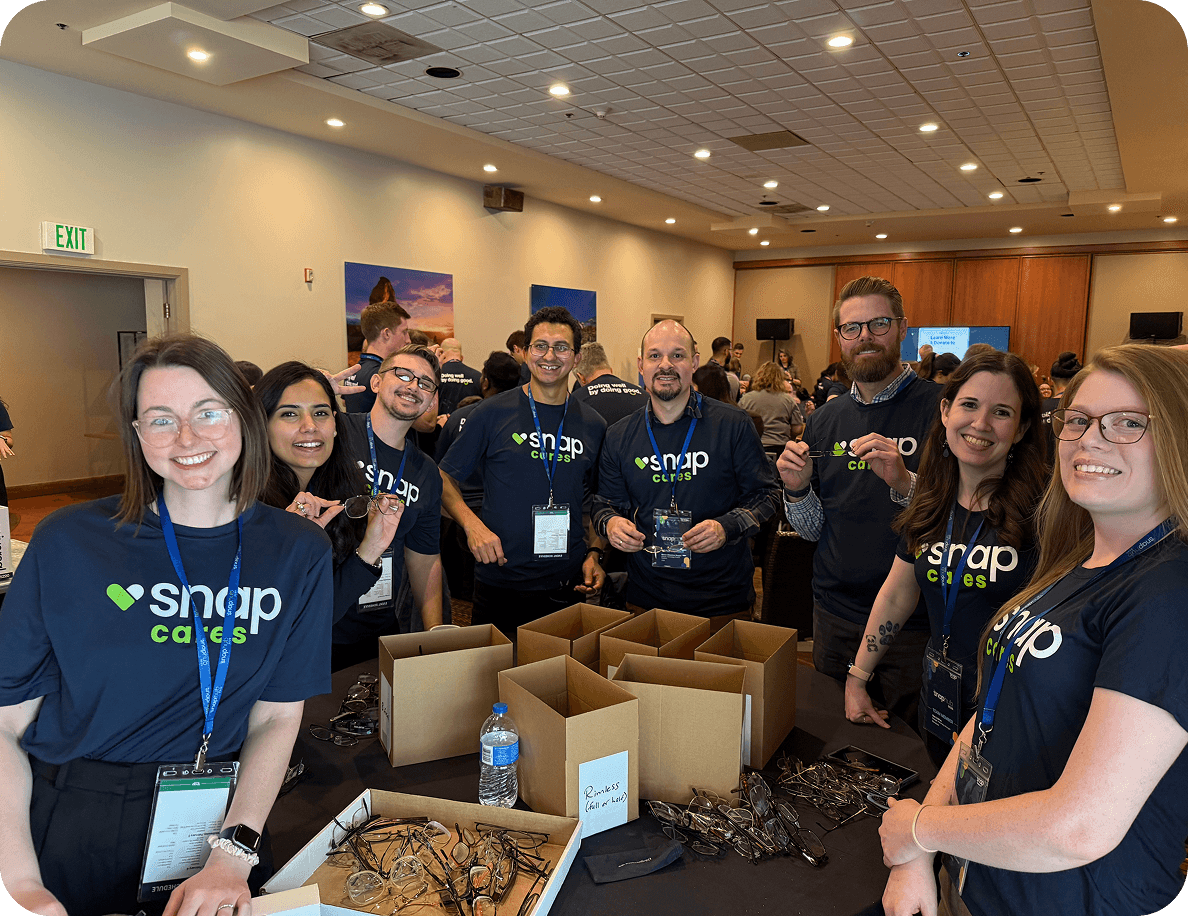 A group of smiling people in "snap cares" shirts stand around a table with boxes and eyeglasses, in a room with many attendees.