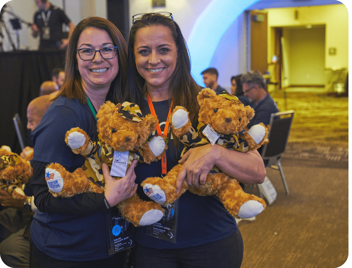 Two women smiling and holding teddy bears in camouflage outfits at an indoor event, with people seated in the background.