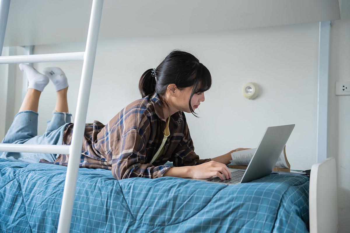 Student lies on blue-quilted dorm bed, feet raised, using laptop and open book; bunk-bed ladder frames scene in minimalist room.