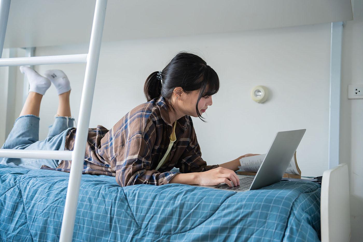 Student lies on blue-quilted dorm bed, feet raised, using laptop and open book; bunk-bed ladder frames scene in minimalist room.