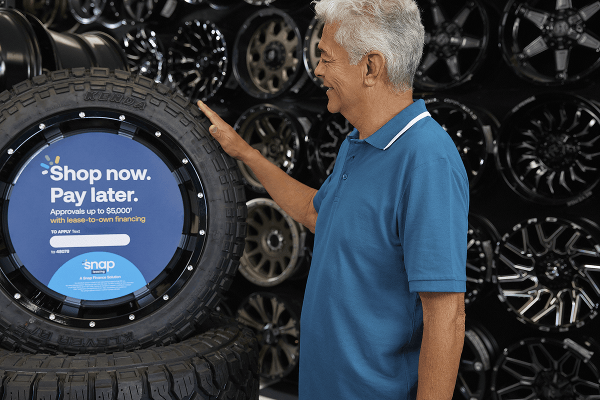 An older man in a blue polo shirt smiles while touching a large tire with a “Shop now. Pay later.” financing sign on display.