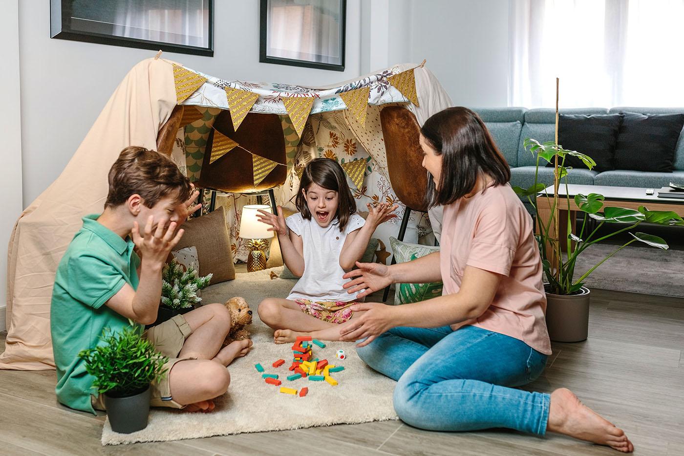 A mother and two children sit on the floor playing with colorful blocks in front of a homemade indoor tent decorated with bunting and pillows.