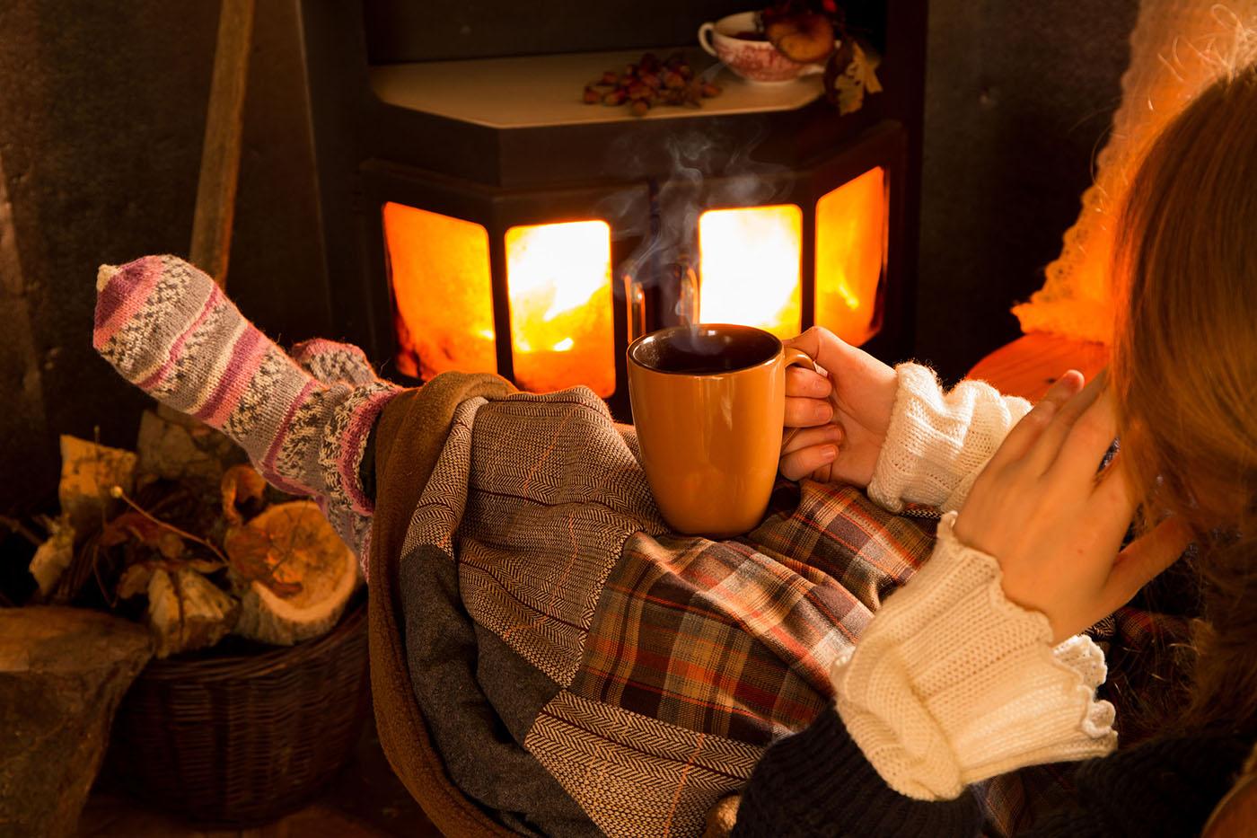 Seated person wrapped in plaid blanket stretches sock-clad feet toward wood stove, holding steaming mug in cozy, firelit living room.