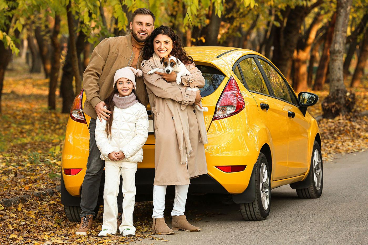 A smiling family with a young girl and a small dog poses in front of a yellow car parked on a leaf-covered road in a wooded area.