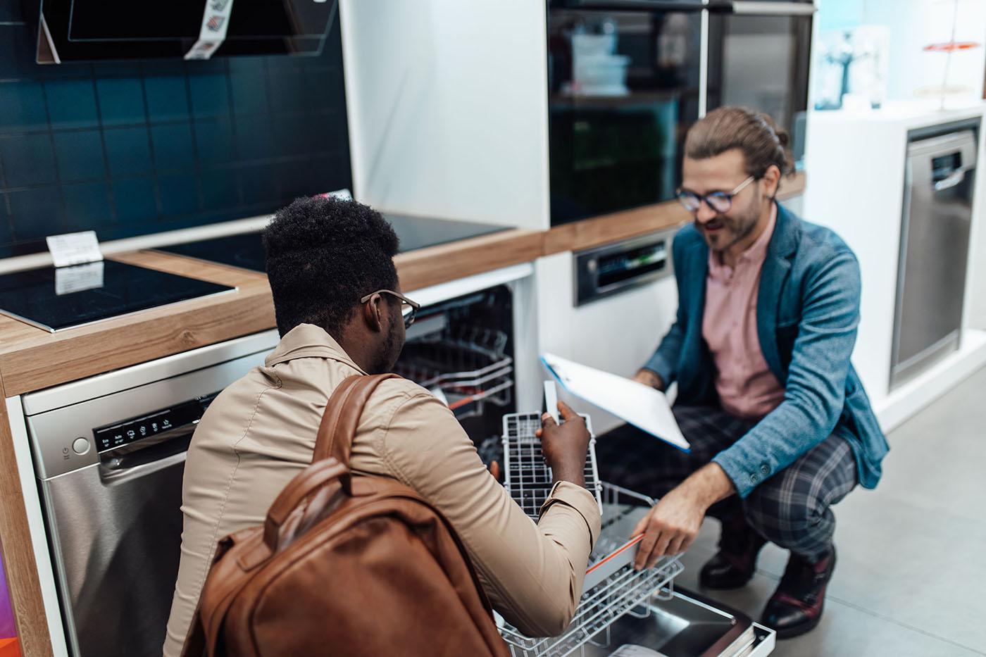 Two men examine an open dishwasher in an appliance store; one is pulling out the rack while the other holds a clipboard, discussing features.