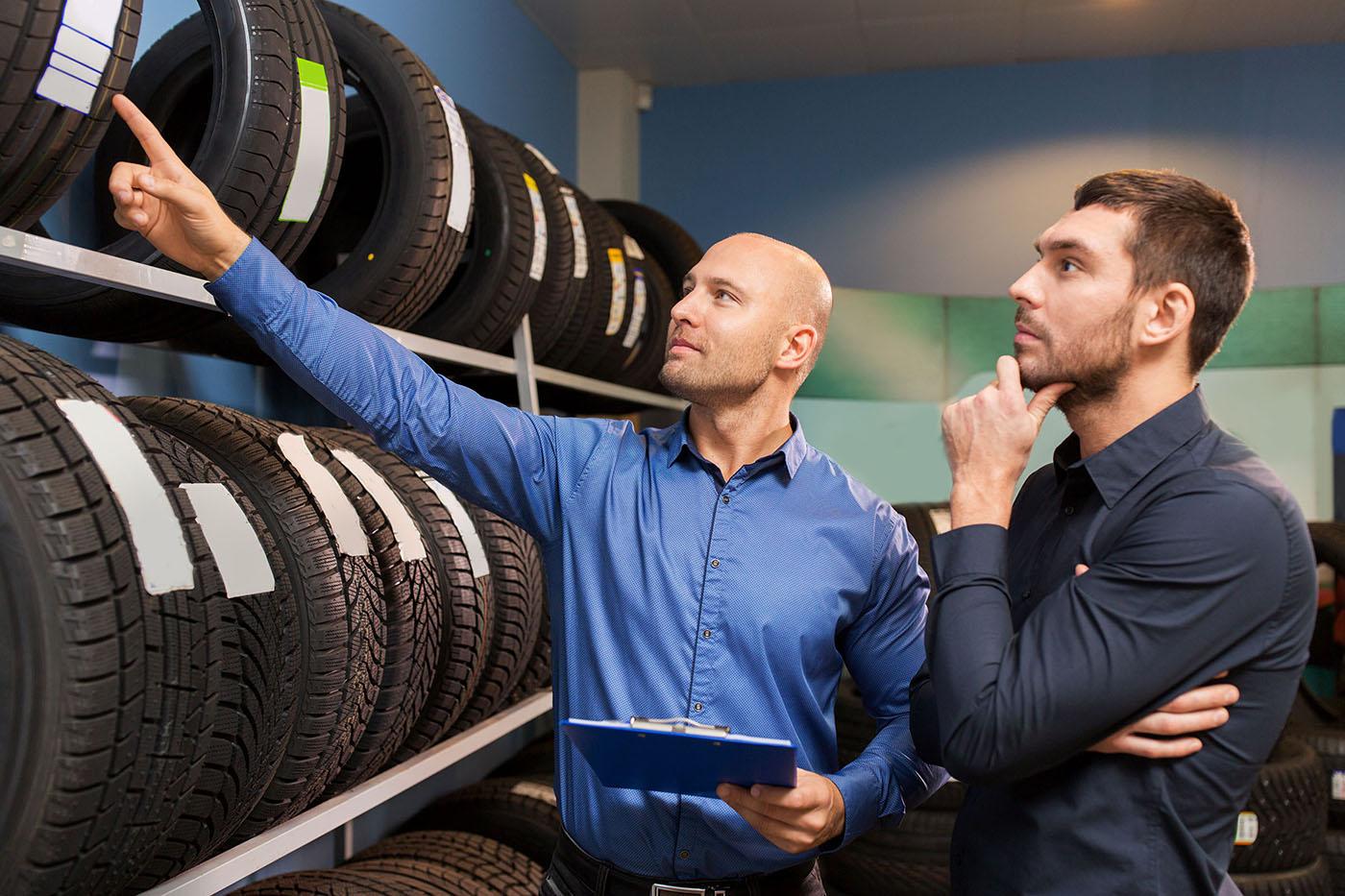 Salesperson holding clipboard points at tire on rack while customer contemplates options among rows of new tires.