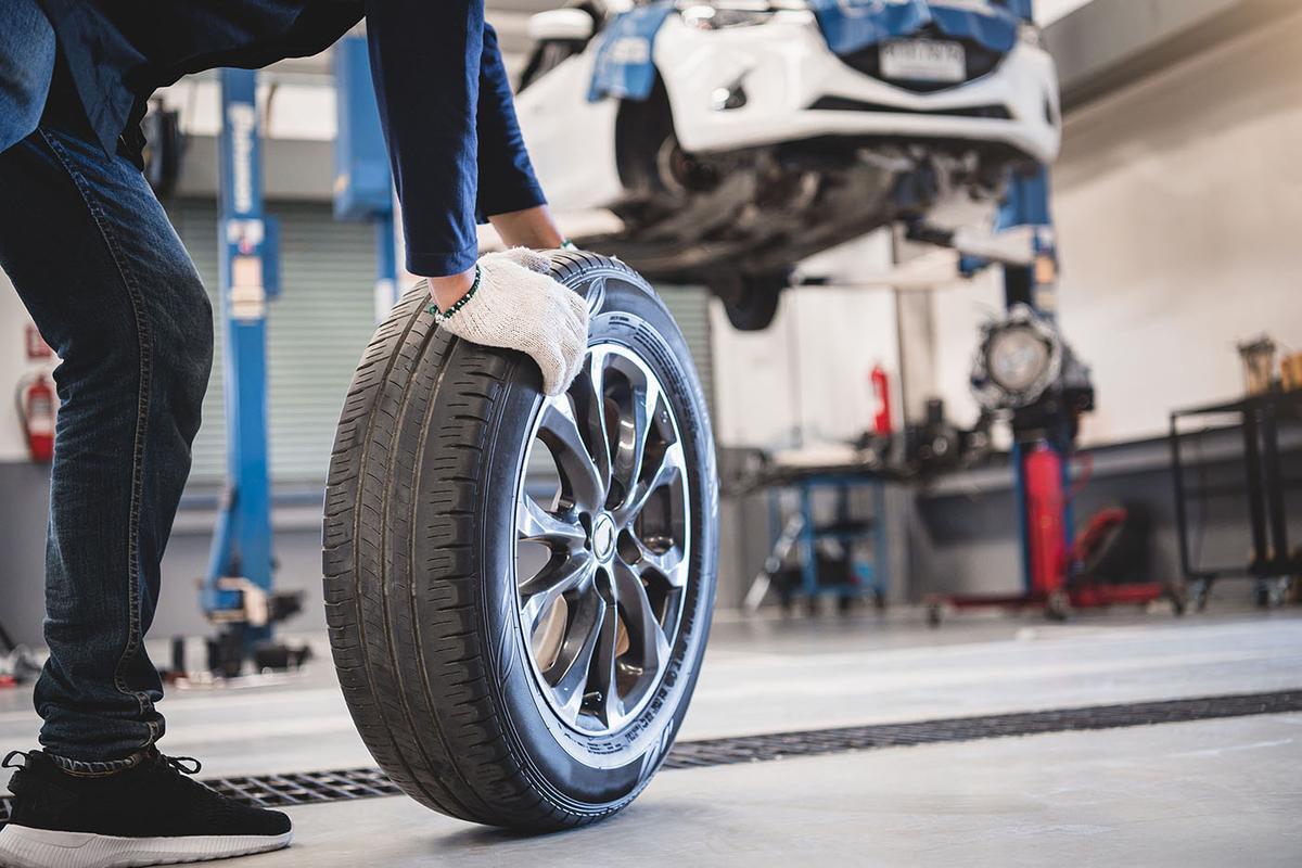 A mechanic wearing gloves rolls a car tire across a garage floor, with a white car lifted on a hydraulic lift in the background.