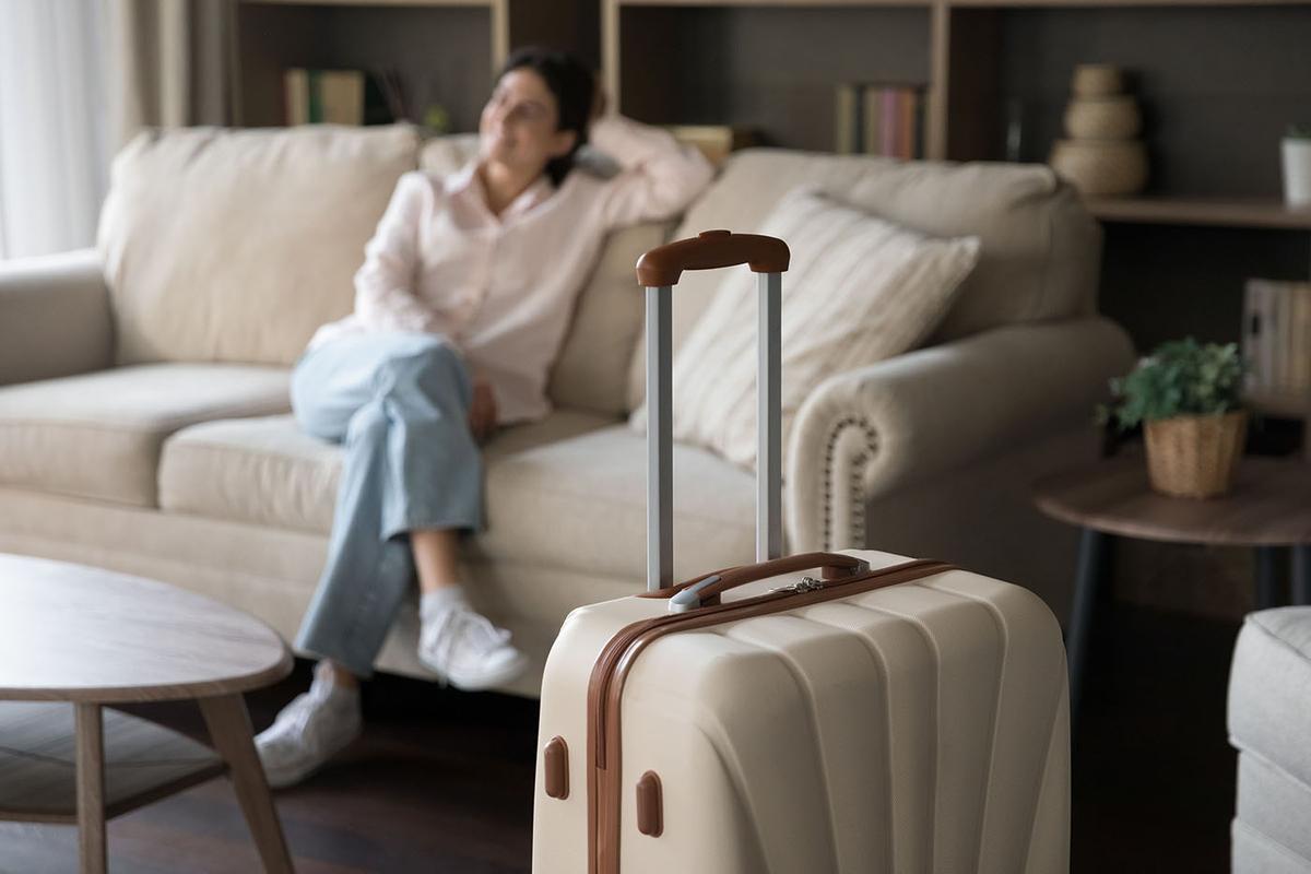 Beige hard-shell suitcase with extended handle stands in foreground; casually dressed woman relaxes on cream sofa in soft-focus background.