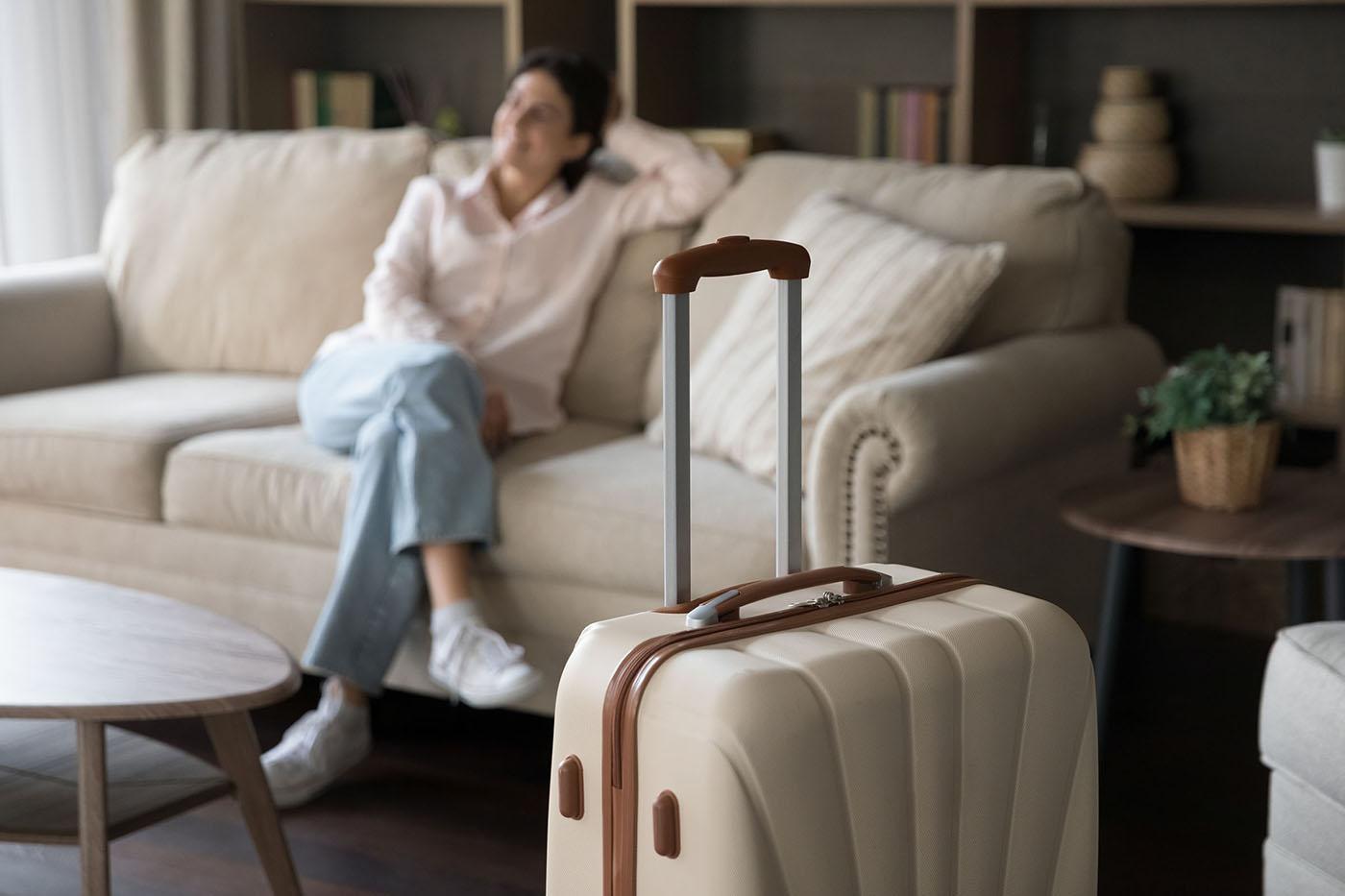 Beige hard-shell suitcase with extended handle stands in foreground; casually dressed woman relaxes on cream sofa in soft-focus background.
