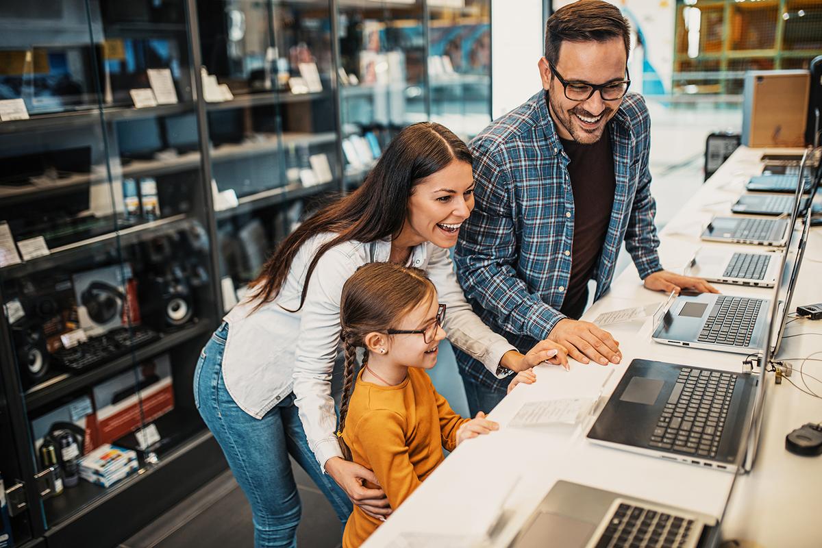 Smiling parents and young daughter lean over a row of open laptops in an electronics store, comparing models and specs.