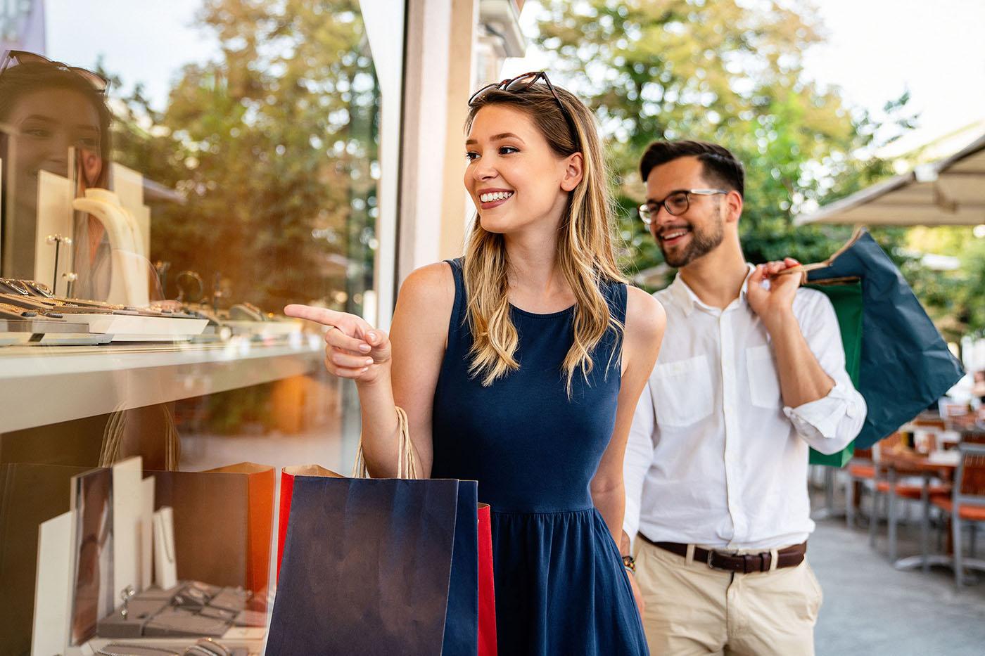 A smiling woman points at jewelry in a store window while holding shopping bags, with a man carrying more bags walking beside her.