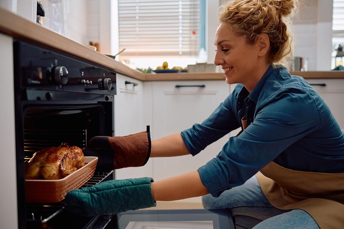A woman wearing oven mitts and an apron smiles as she places a roasted chicken in a baking dish into a modern kitchen oven.