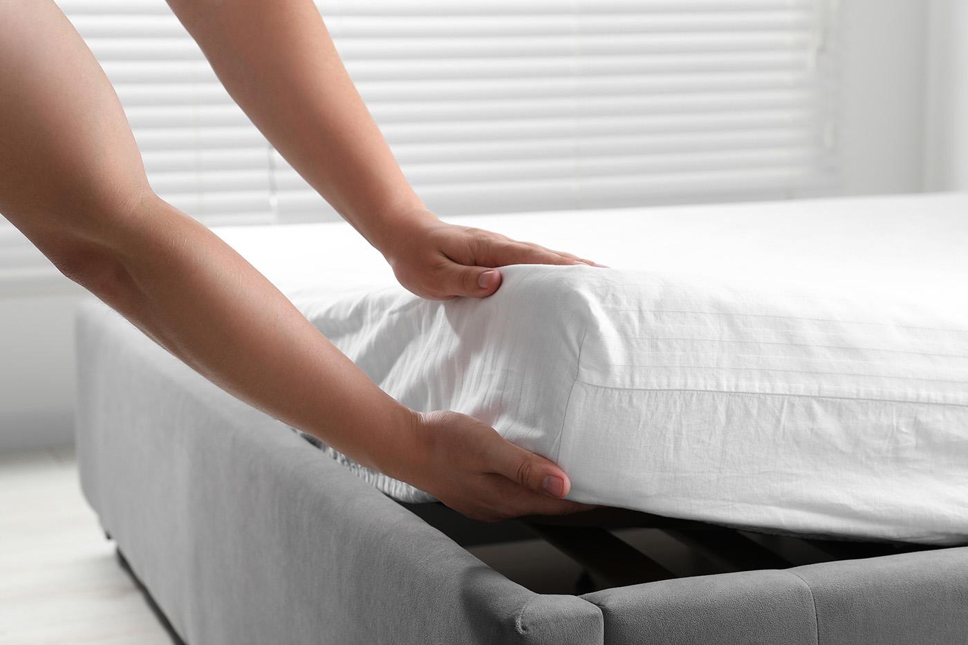 Close-up of a person’s hands putting a white fitted sheet onto a mattress on a gray upholstered bed frame.