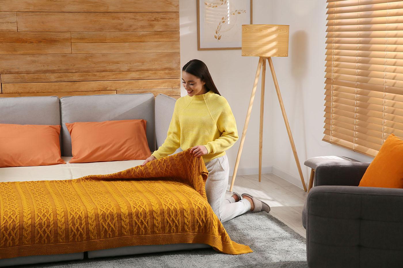 A woman in a yellow sweater kneels beside a bed, neatly spreading a textured orange blanket in a cozy, sunlit bedroom.