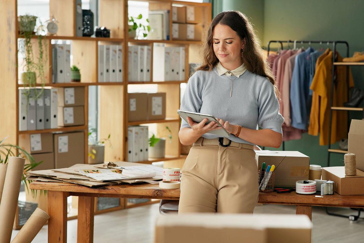 A woman stands in a small business workspace, holding a tablet and smiling, surrounded by boxes, packaging supplies, and clothing on racks.