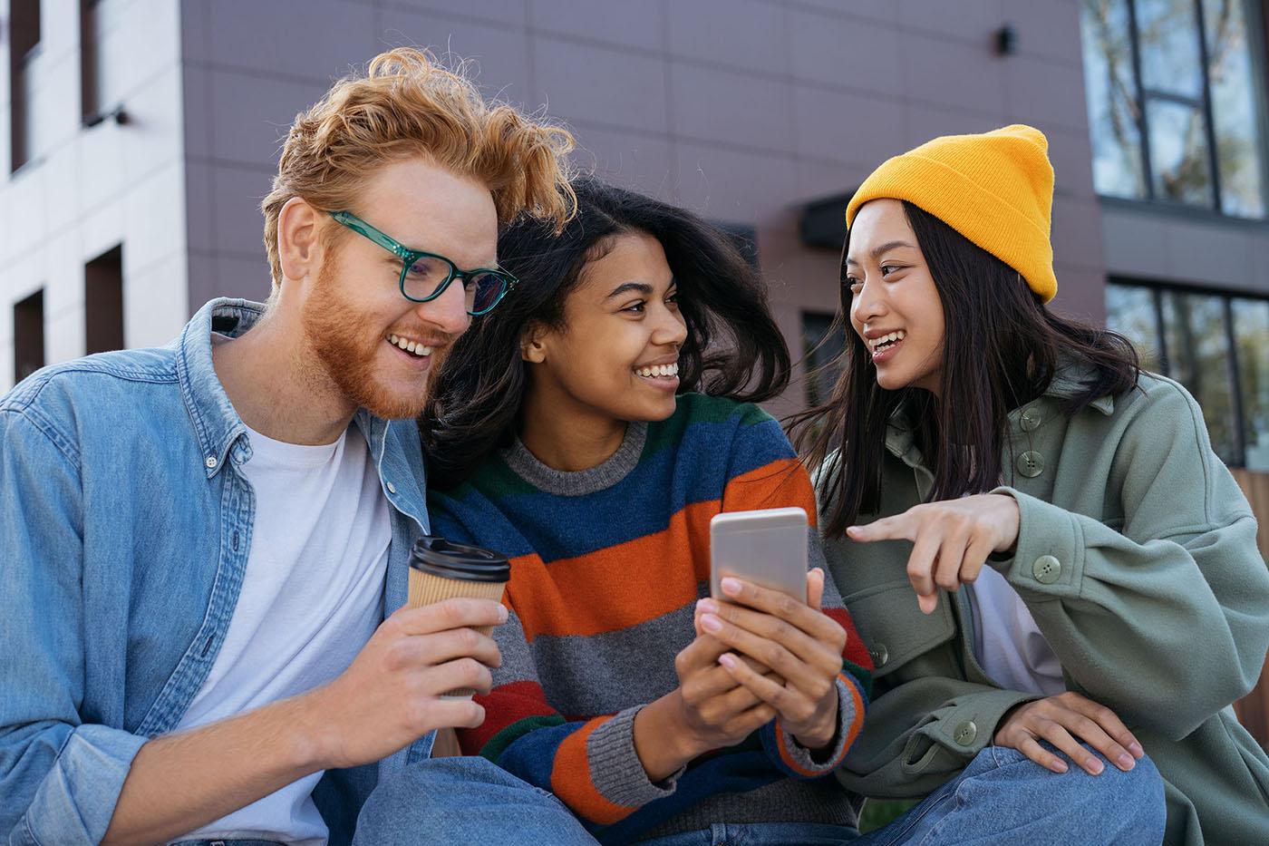 Three friends sit outside a modern building, smiling and laughing while looking at something on a smartphone; one holds a coffee cup, another points.