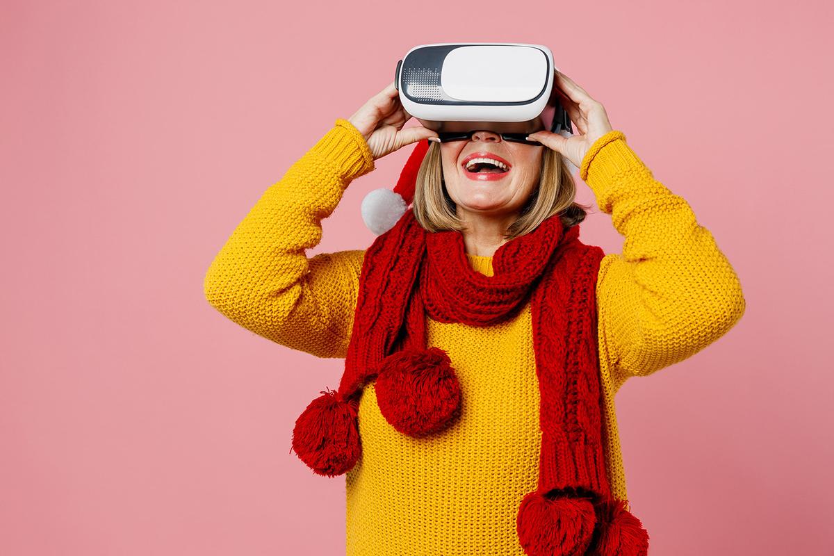 A cheerful woman in a red scarf and Santa hat wears a VR headset, smiling while experiencing virtual reality against a pink background.