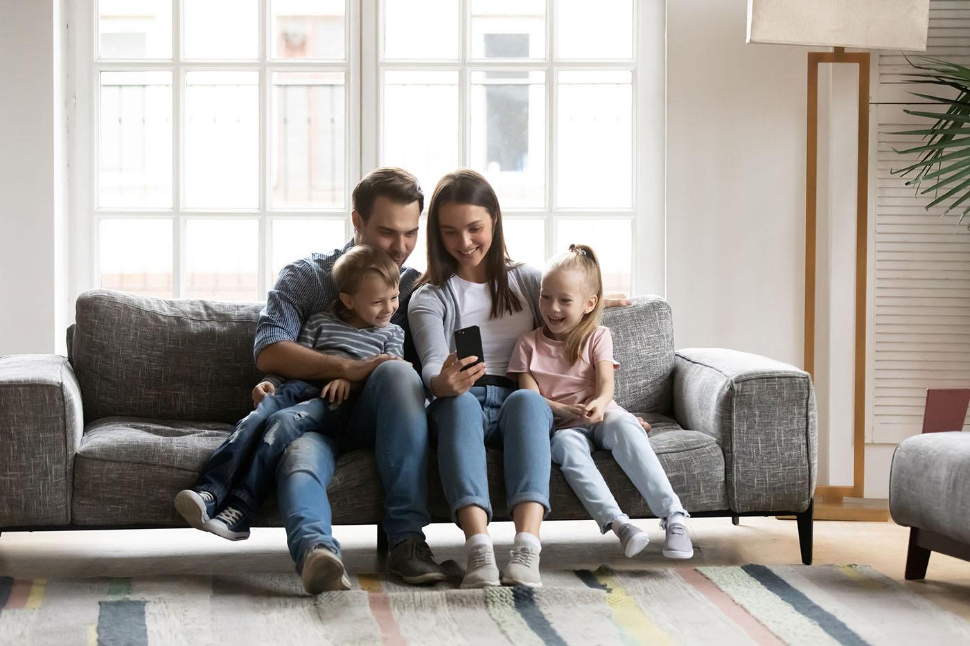 A smiling family of four sits on a gray sofa, looking at a smartphone together in a bright living room with large windows.