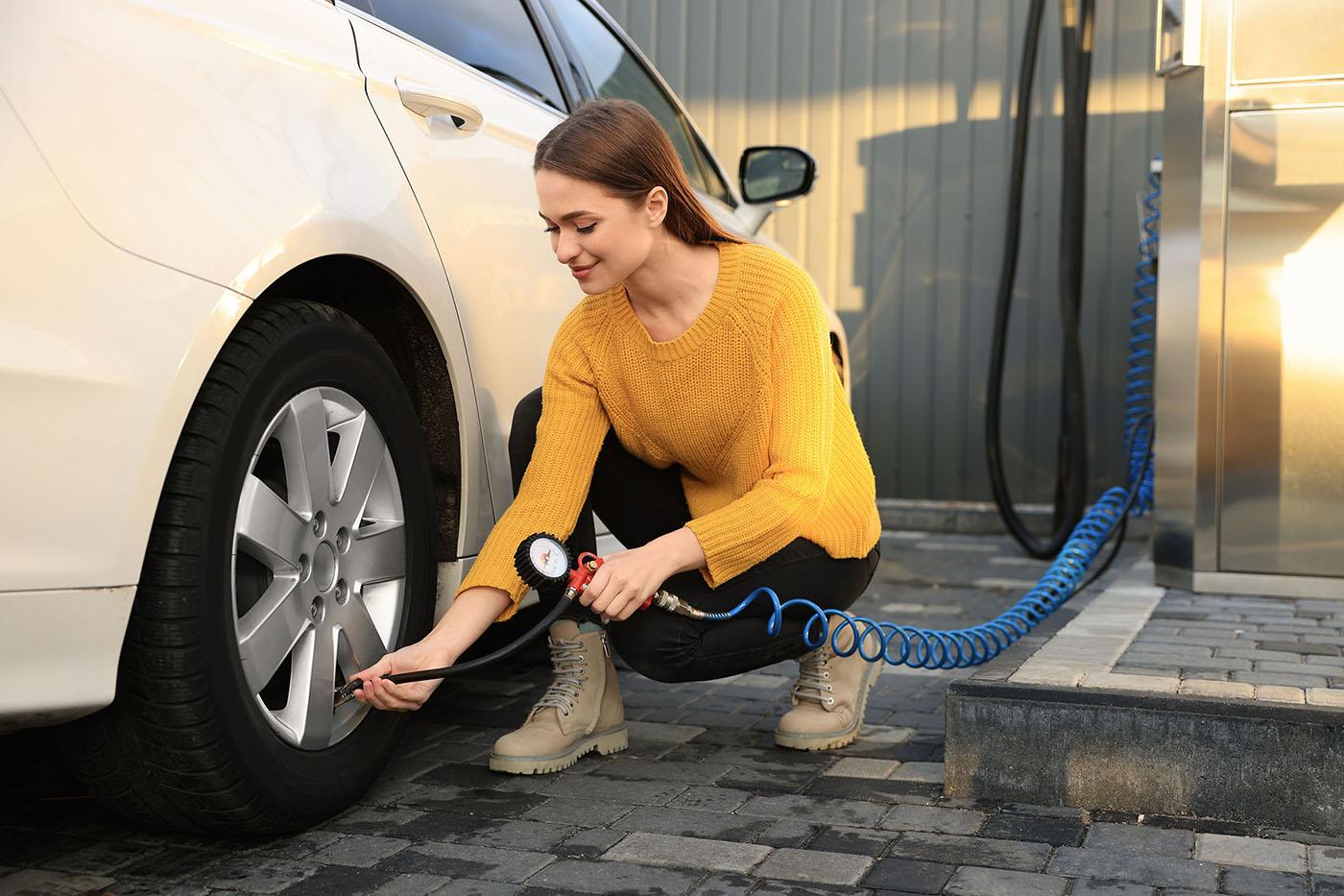 A woman in a yellow sweater crouches beside a car, using an air hose with a pressure gauge to inflate the front tire at a service station.