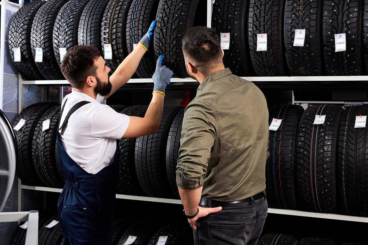 A tire shop worker in overalls explains tire options to a customer, pointing at a row of winter tires on a display rack.