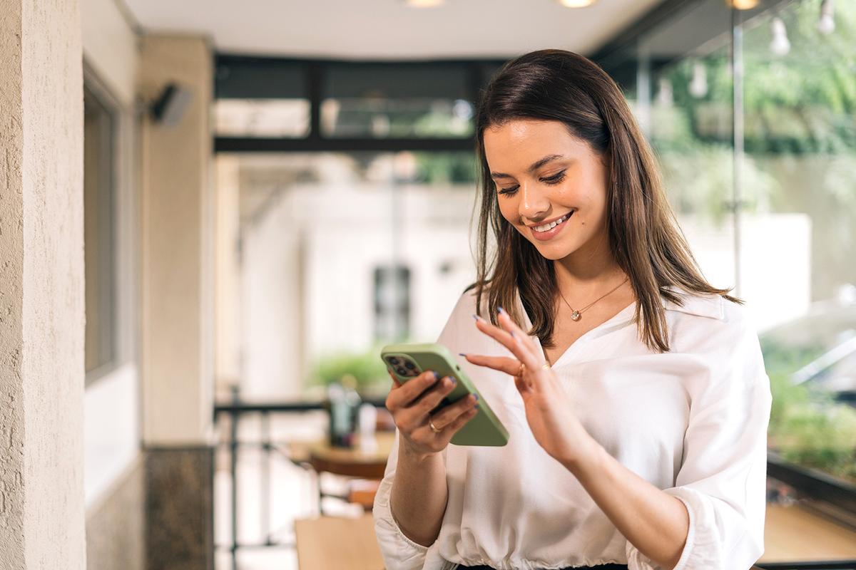 A woman in a white blouse stands indoors near a window, smiling while using a green smartphone with one hand.