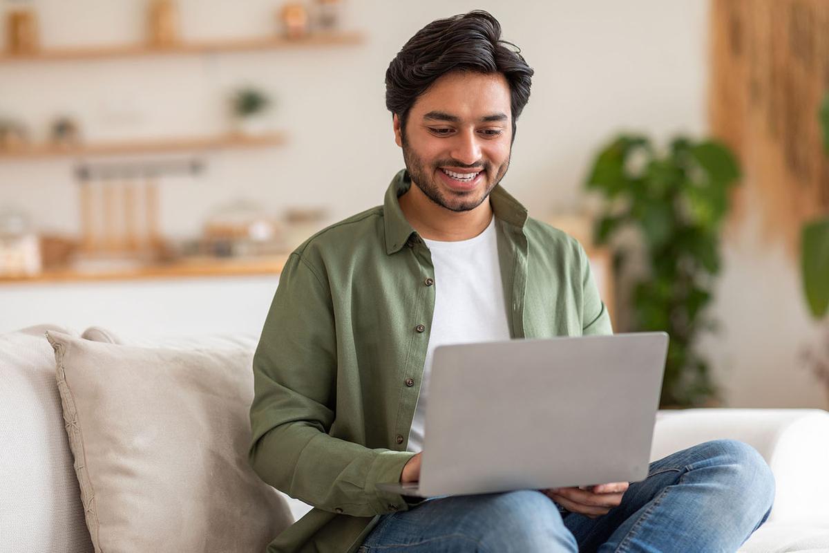 A smiling man in a green shirt sits on a couch using a laptop in a bright, cozy living room with plants and shelves in the background.
