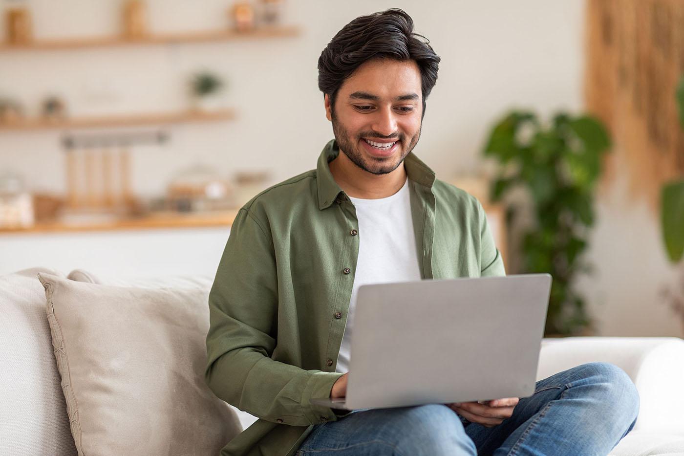 A smiling man in a green shirt sits on a couch using a laptop in a bright, cozy living room with plants and shelves in the background.