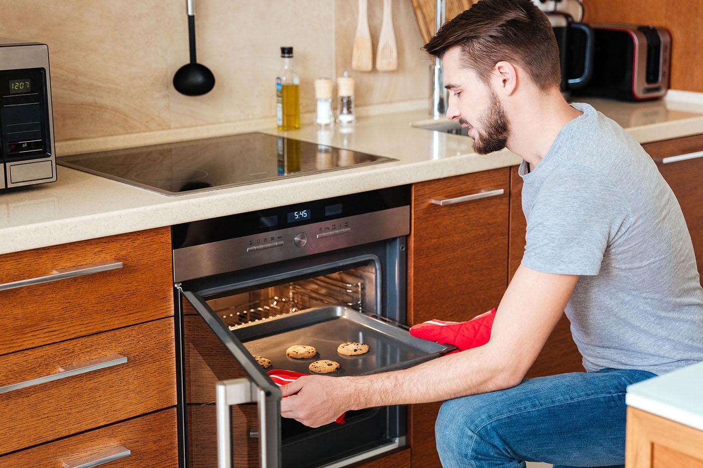 A man wearing red oven mitts removes a baking tray with chocolate chip cookies from a modern kitchen oven.
