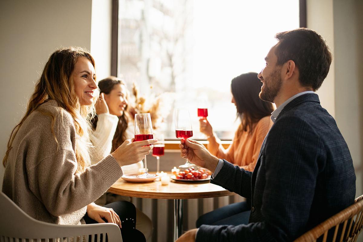 Two couples sit at a round table by a window, smiling and clinking glasses of red wine in a cozy, warmly lit setting.