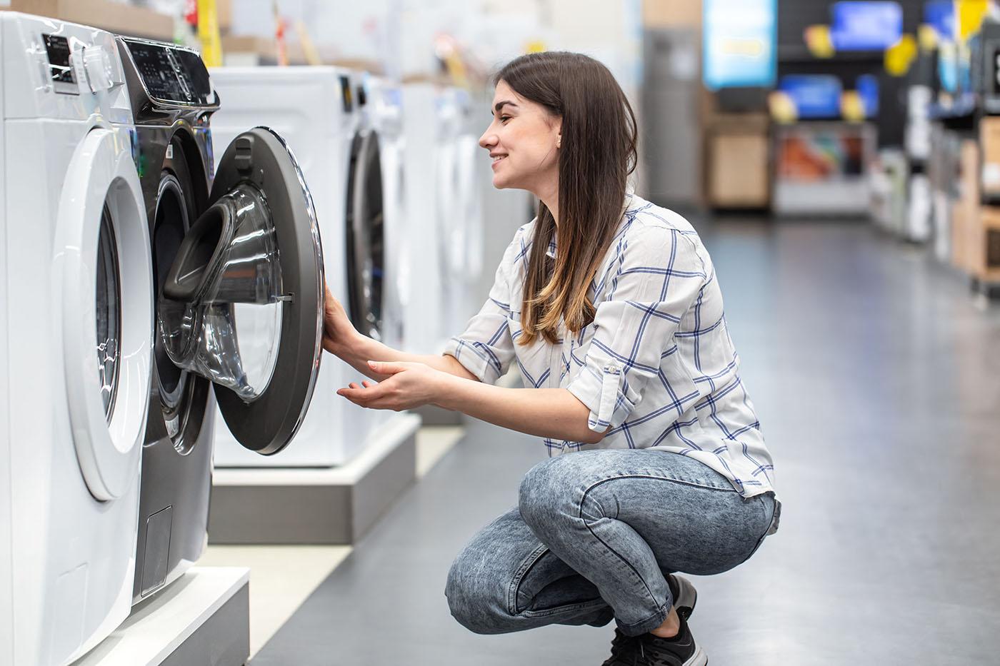 A woman in a plaid shirt crouches to inspect the open door of a front-loading washing machine in an appliance store.