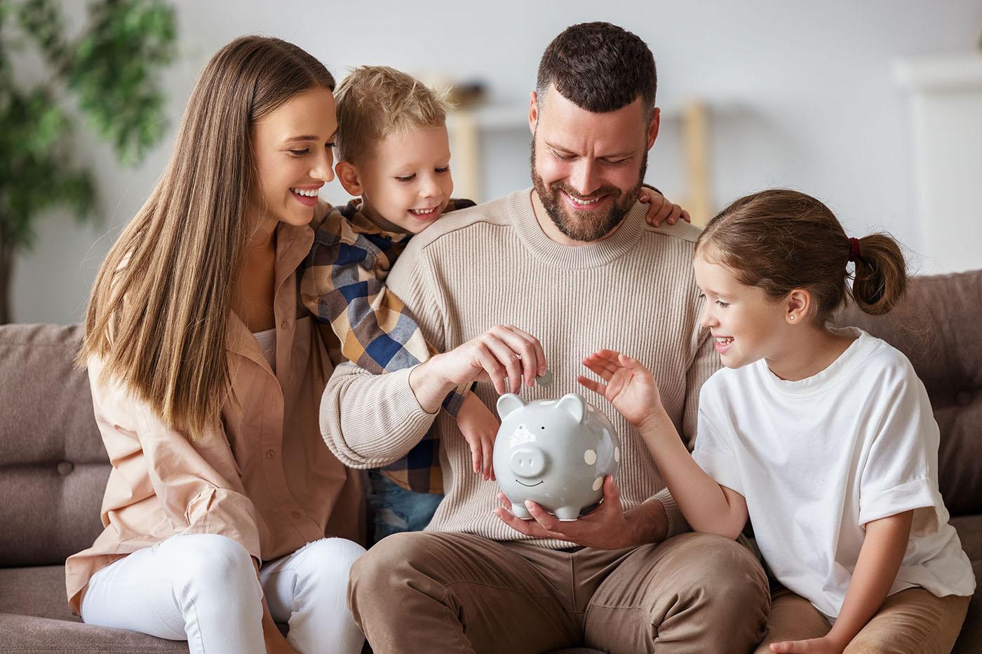 A smiling family of four sits on a couch, placing coins into a gray polka-dotted piggy bank held by the father, promoting saving together.