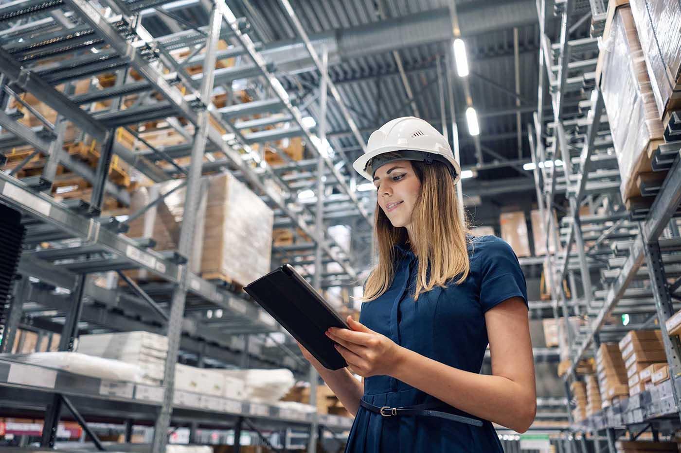 A woman wearing a hard hat and navy dress holds a tablet while inspecting inventory in a large warehouse with tall metal shelving.