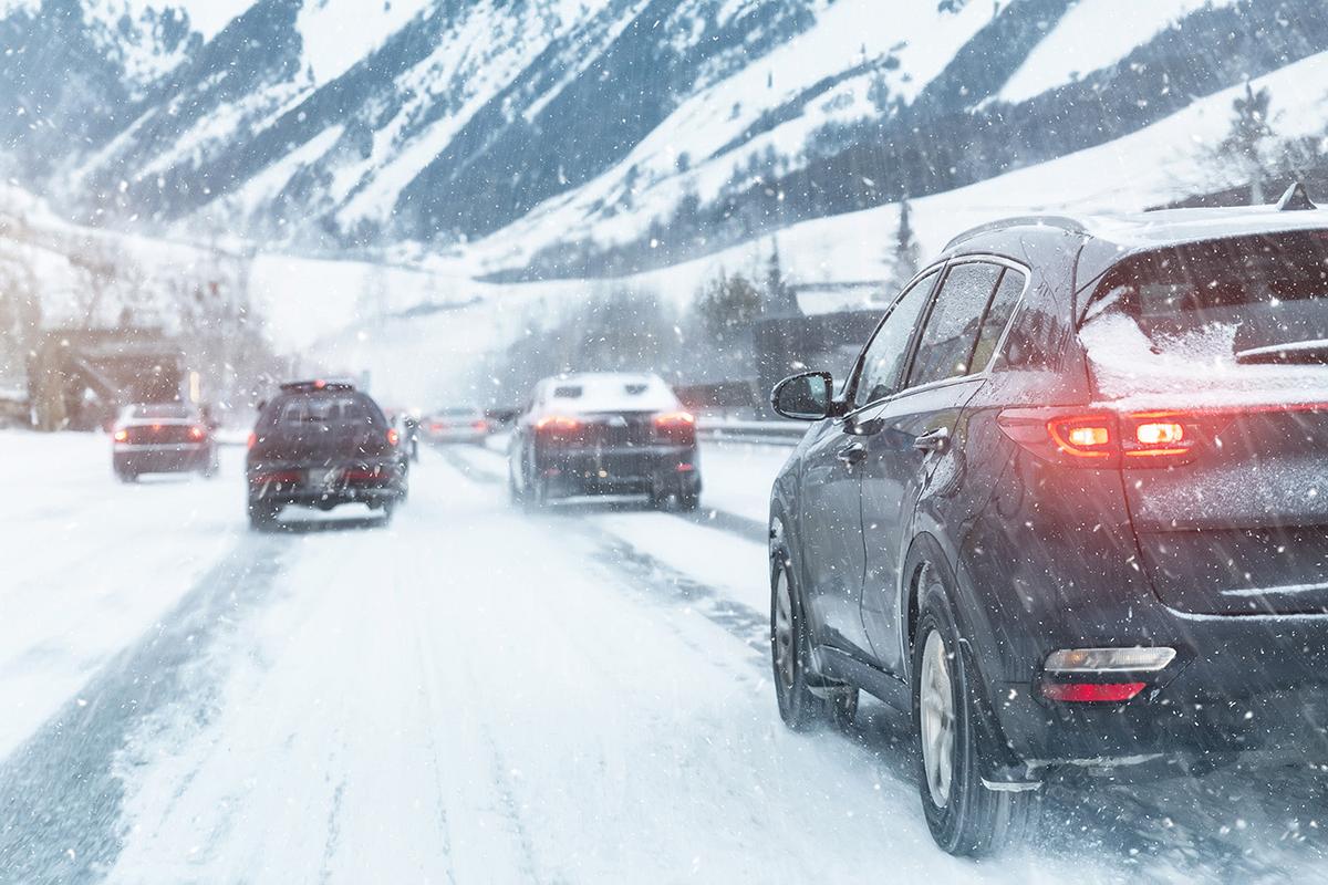 Cars drive cautiously on a snow-covered mountain road during a snowfall, with brake lights illuminated and snow accumulation visible on vehicles.