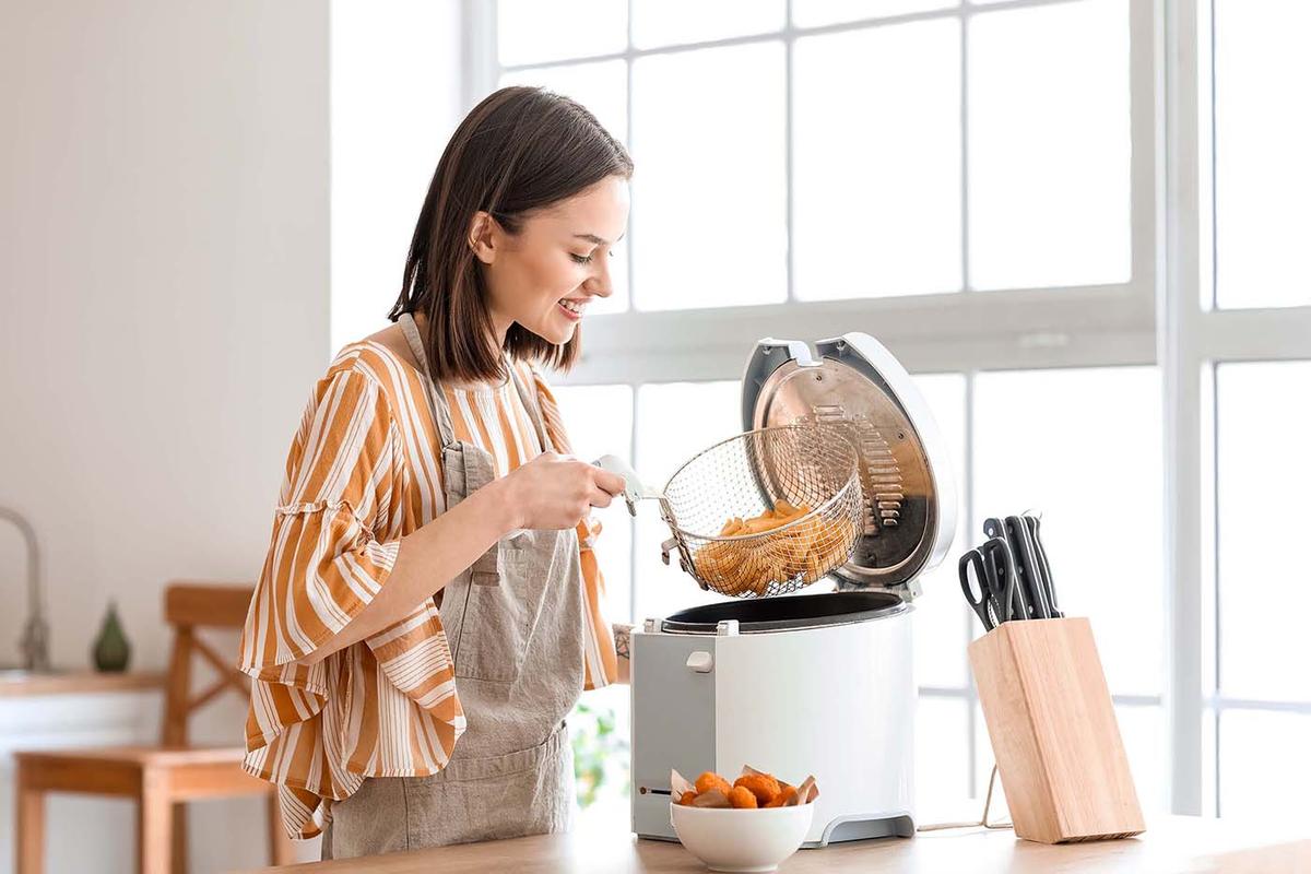 A woman in an apron smiles while lifting a fryer basket filled with cooked food from an electric deep fryer in a bright kitchen.