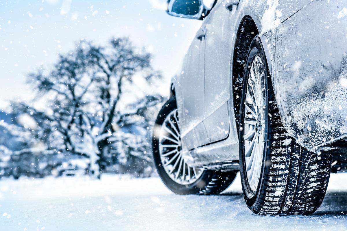 Low-angle view of car with snow-coated tires driving through falling snow on icy road; blurred snow-laden tree in background.