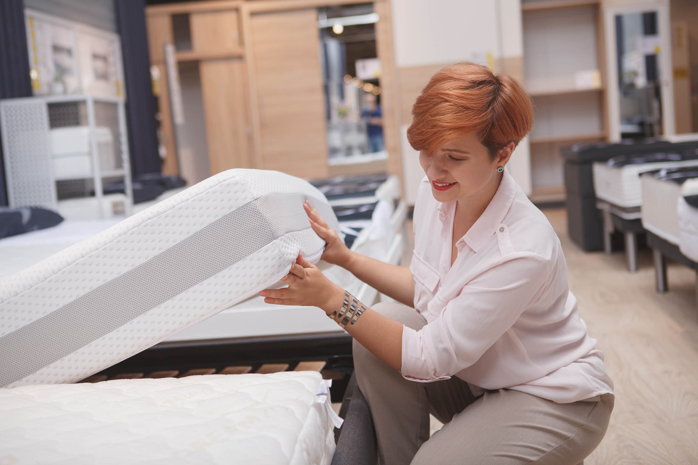 A woman with short hair inspects a white mattress in a store, smiling and examining its quality.