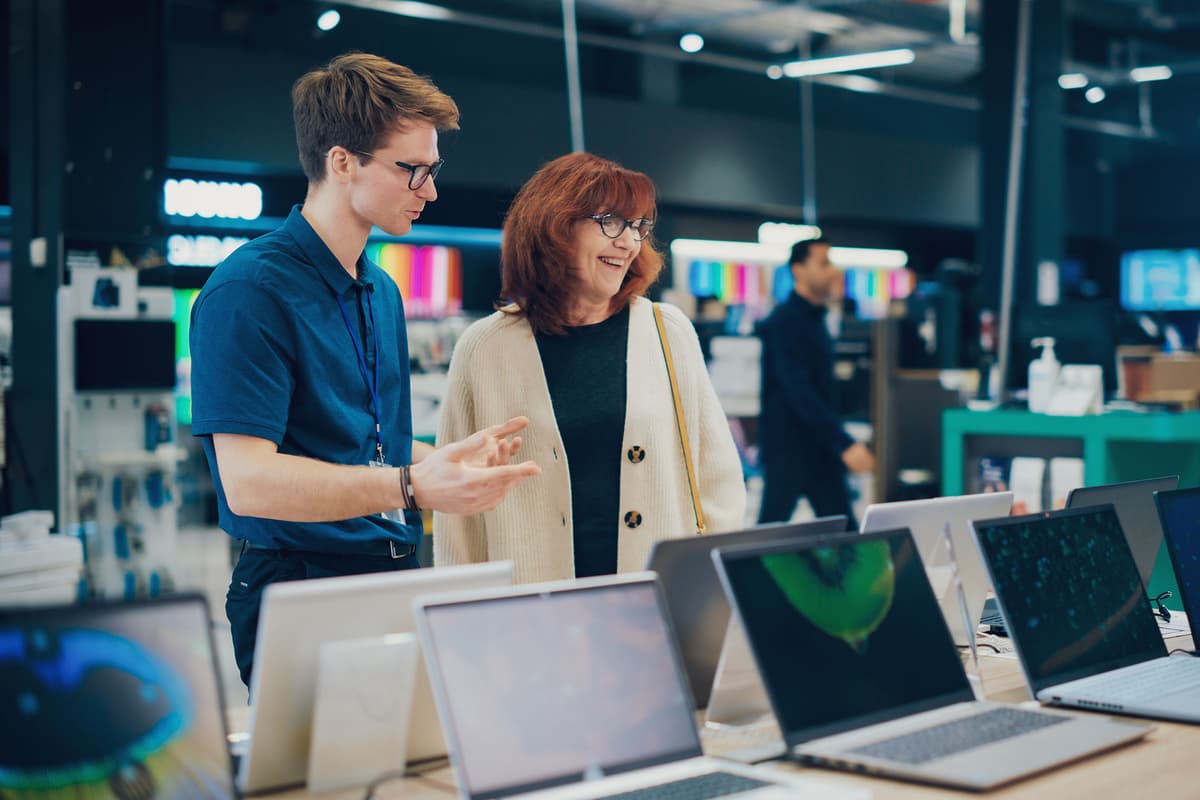 A store employee assists a smiling customer in choosing a laptop from a display in an electronics store.