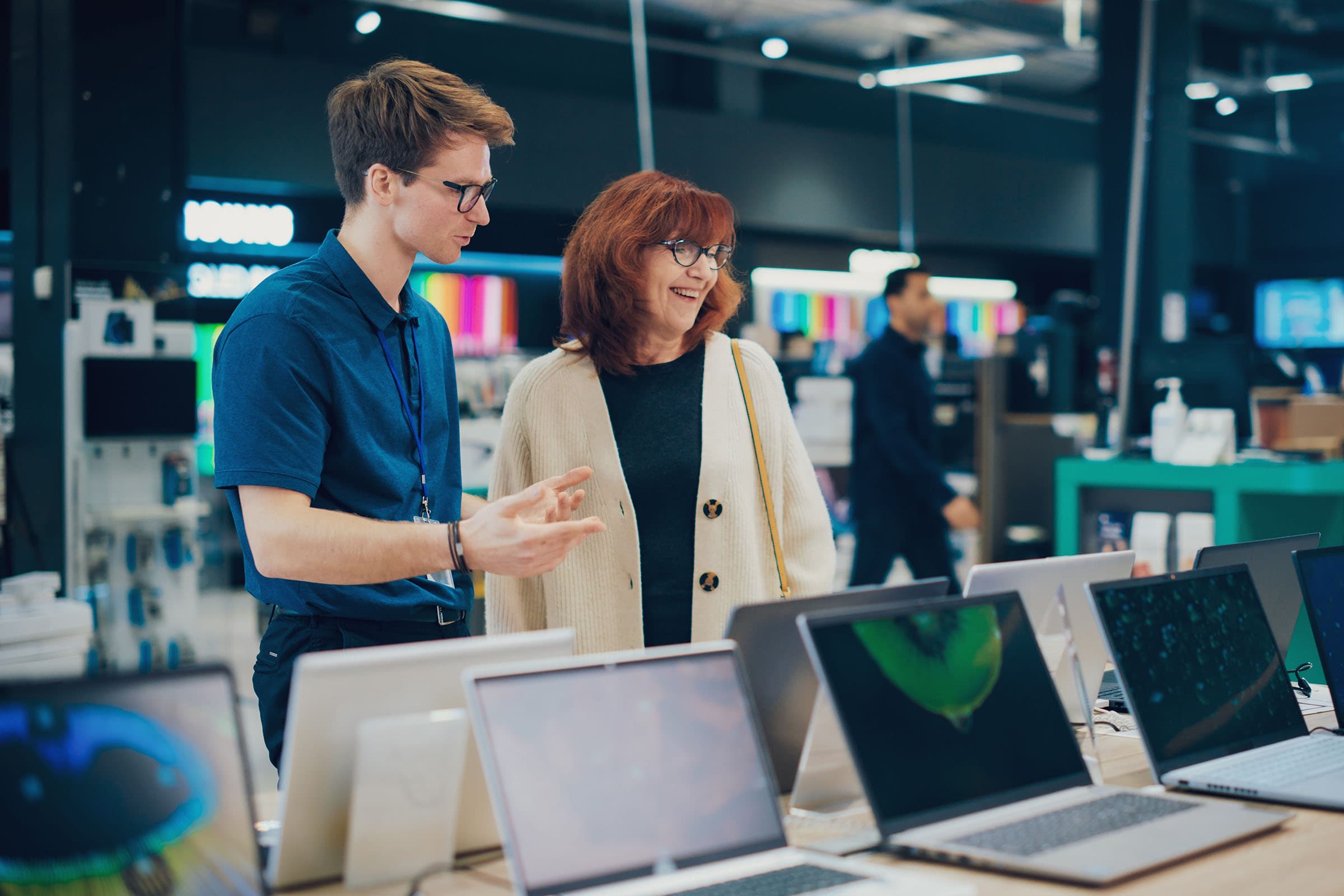 A store employee assists a smiling customer in choosing a laptop from a display in an electronics store.
