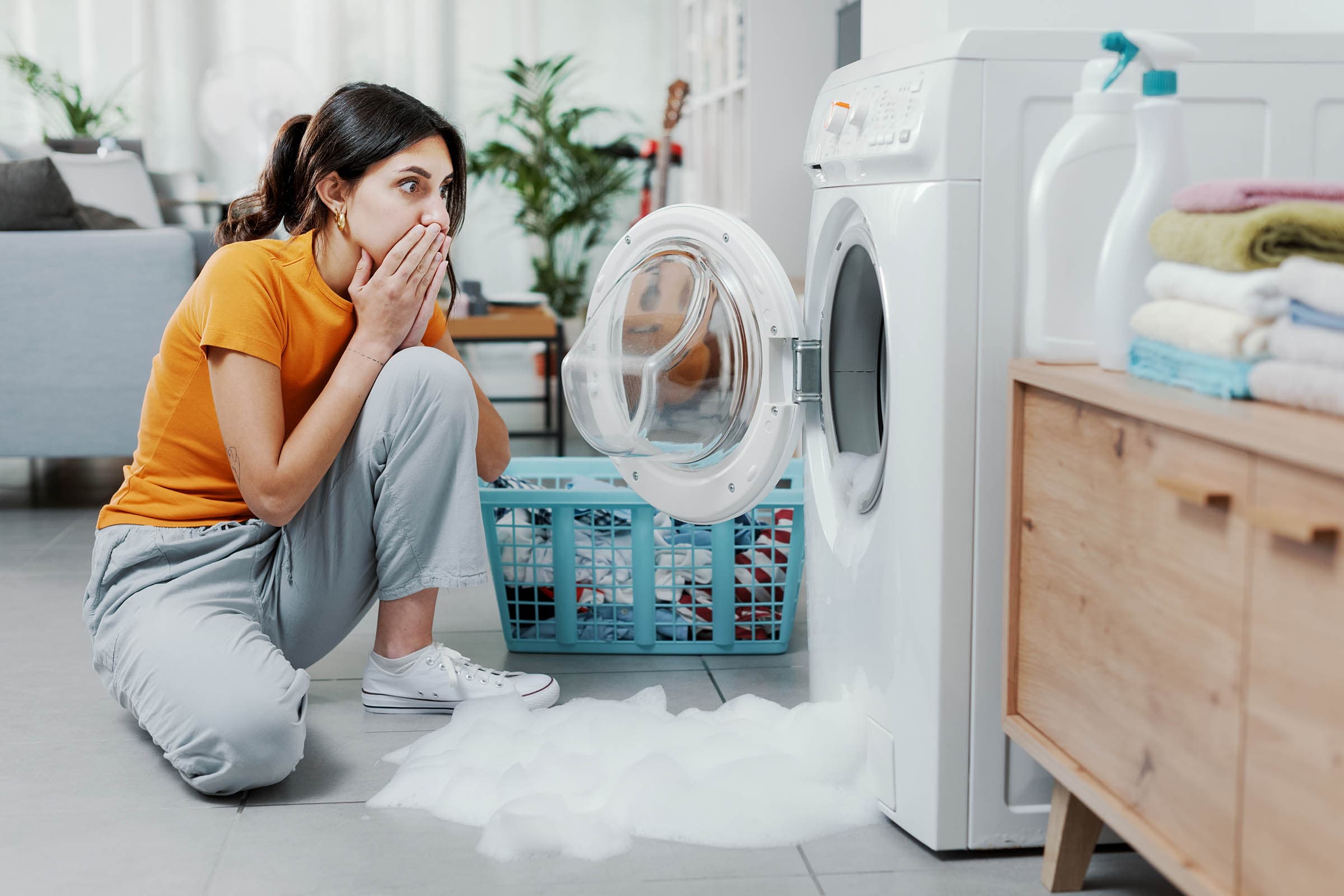A woman in an orange shirt kneels, shocked, in front of a washing machine with soap suds overflowing onto the floor.