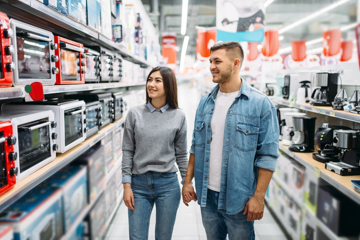 A smiling couple holds hands while shopping for kitchen appliances in a store, surrounded by shelves of toasters and kitchen gadgets.