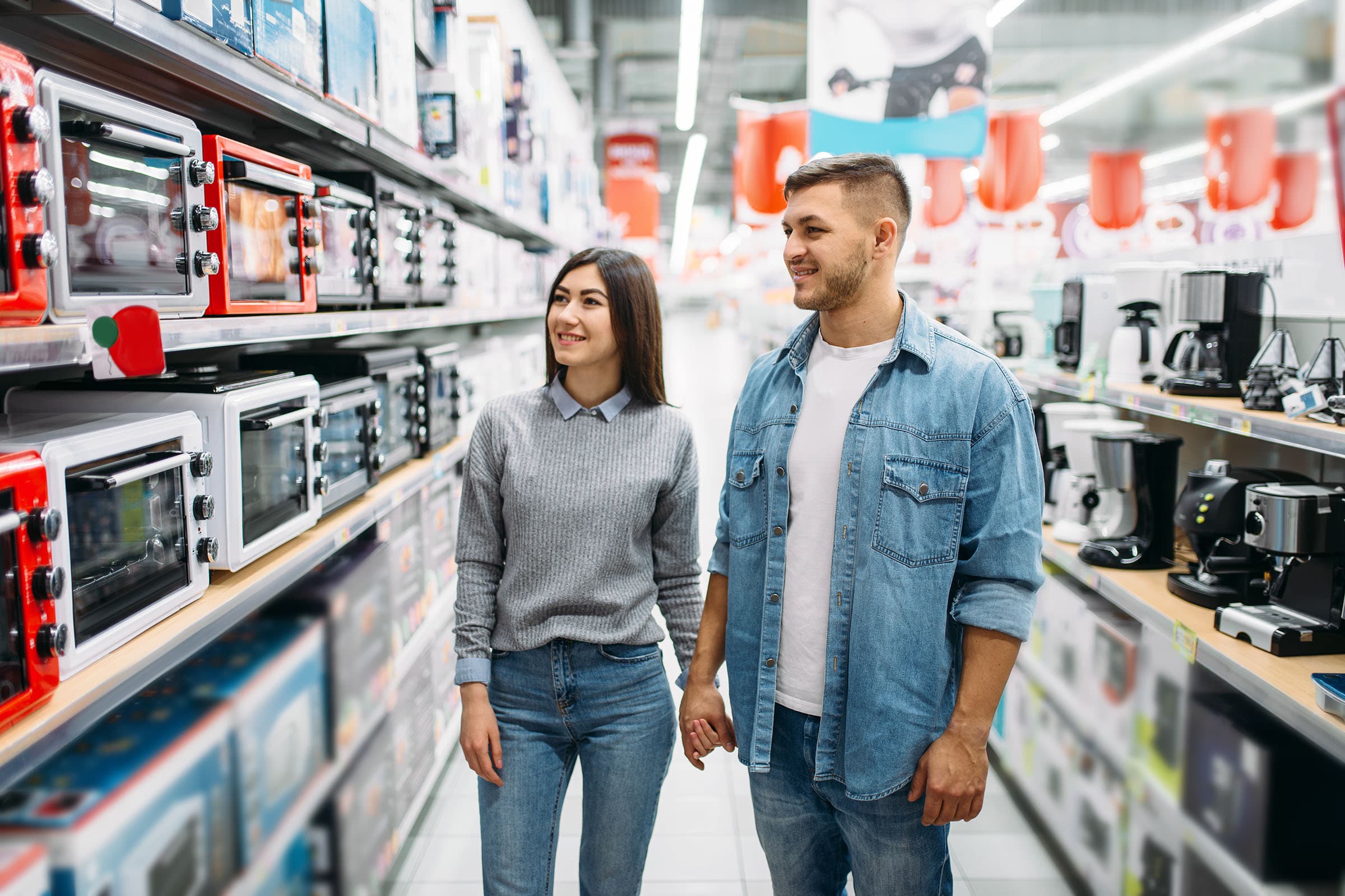 A smiling couple holds hands while shopping for kitchen appliances in a store, surrounded by shelves of toasters and kitchen gadgets.