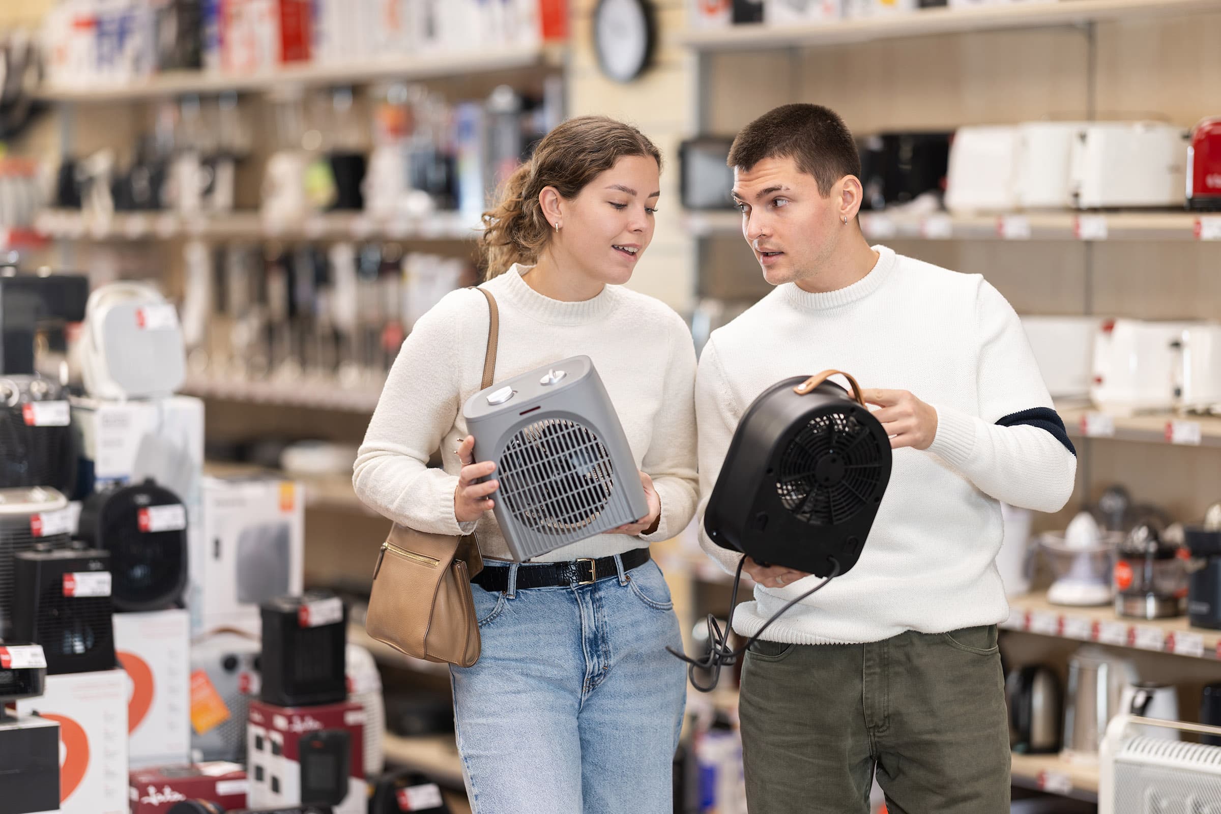 Two people in a store discussing portable heaters, holding a gray and black model, with shelves of appliances in the background.