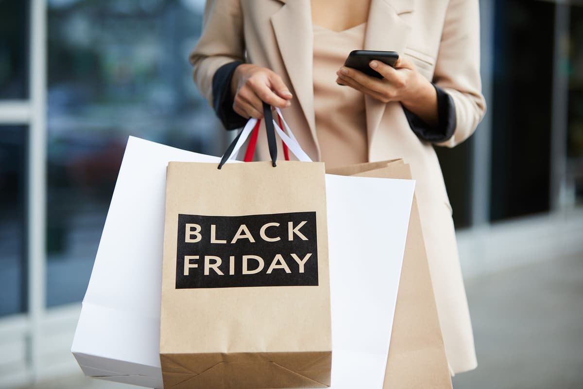 Person holding Black Friday shopping bags and a smartphone, dressed in a beige coat, standing outside a store.