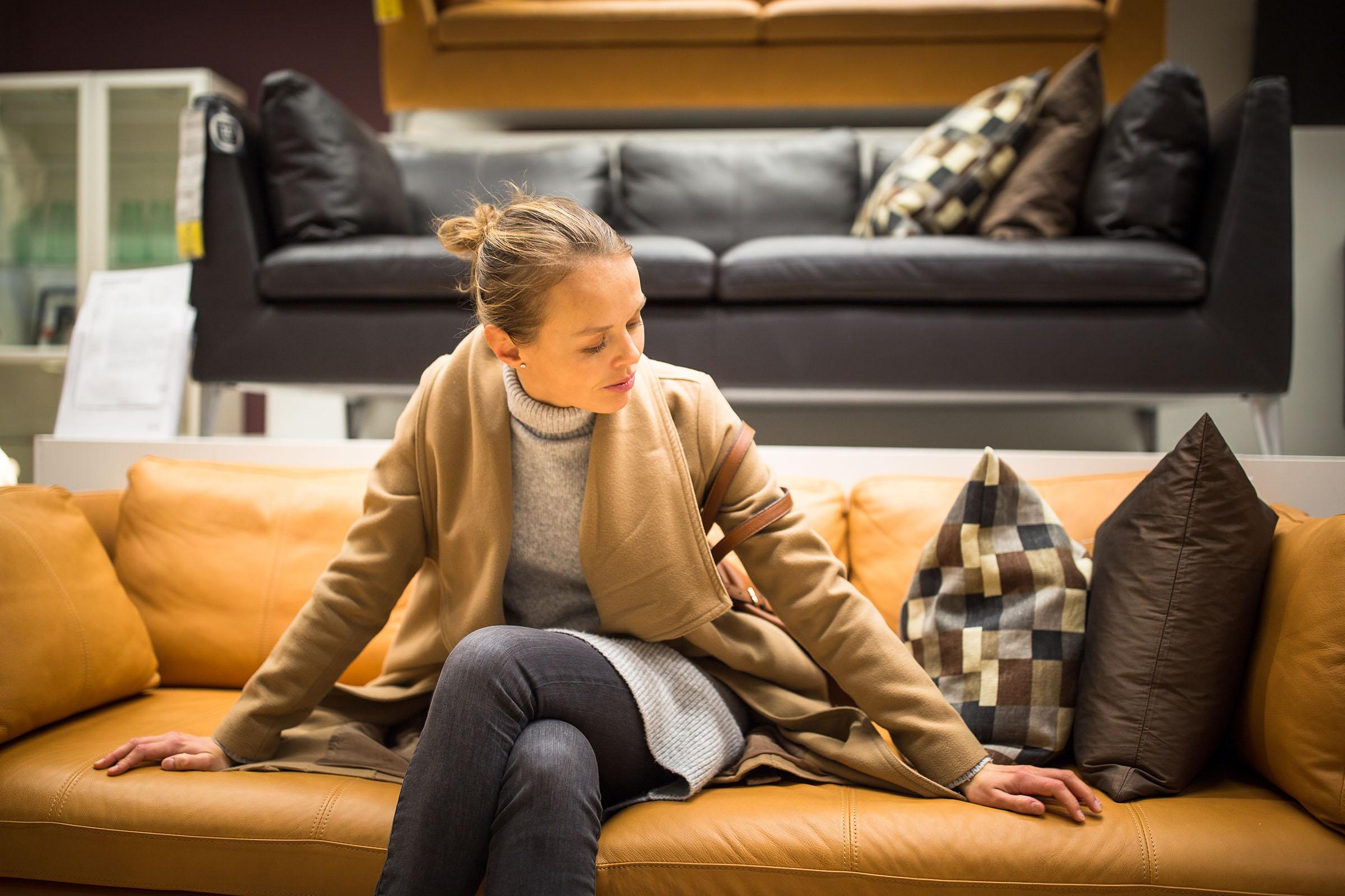 Woman in tan coat sits on tan leather sofa in showroom, testing cushion firmness near two patterned throw pillows.