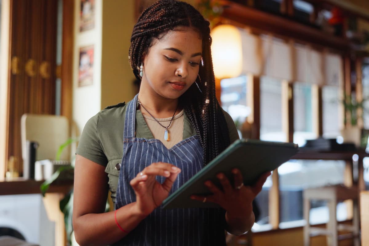A woman in a striped apron uses a tablet in a cozy cafe setting.