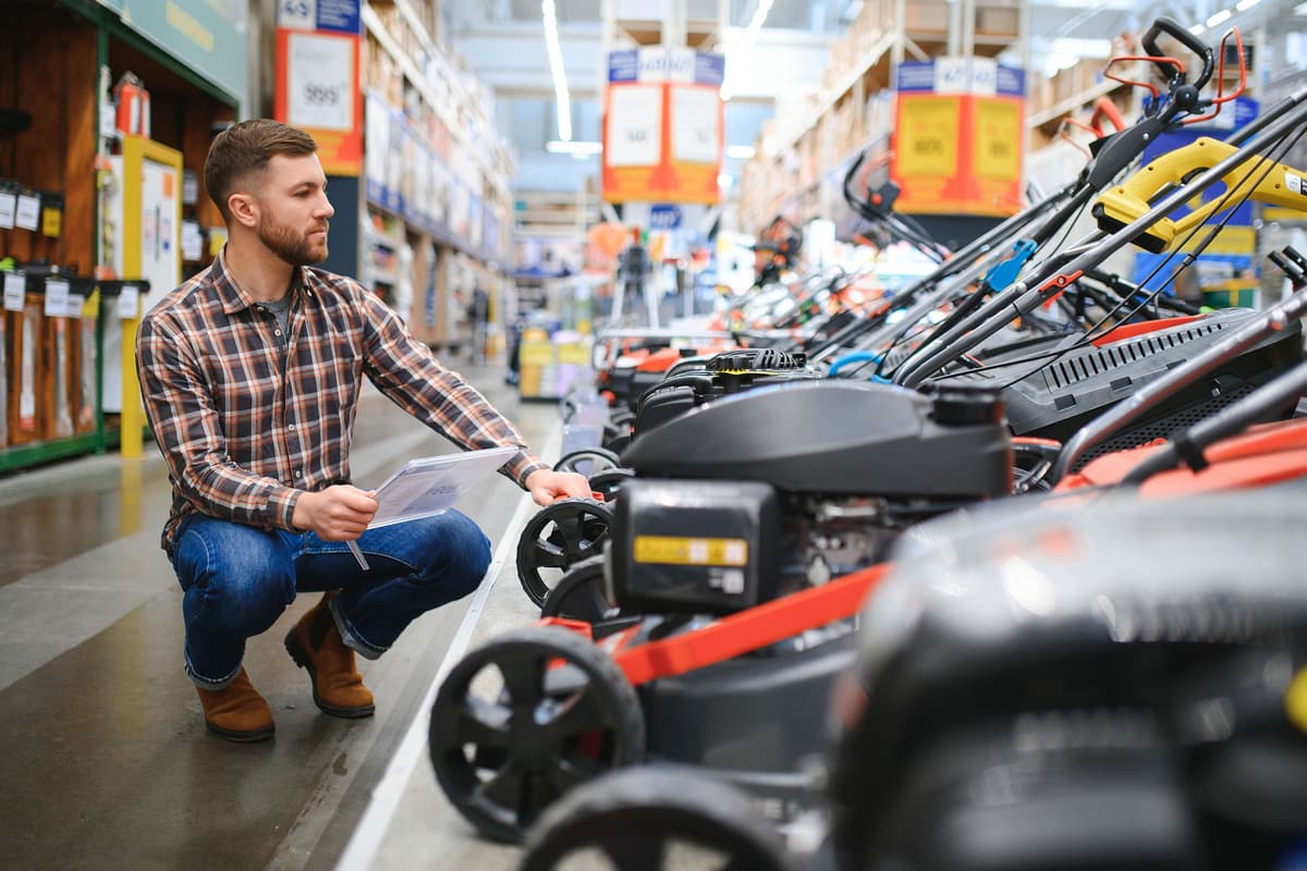 Man in a hardware store kneeling and examining a row of lawn mowers while holding product information sheets.