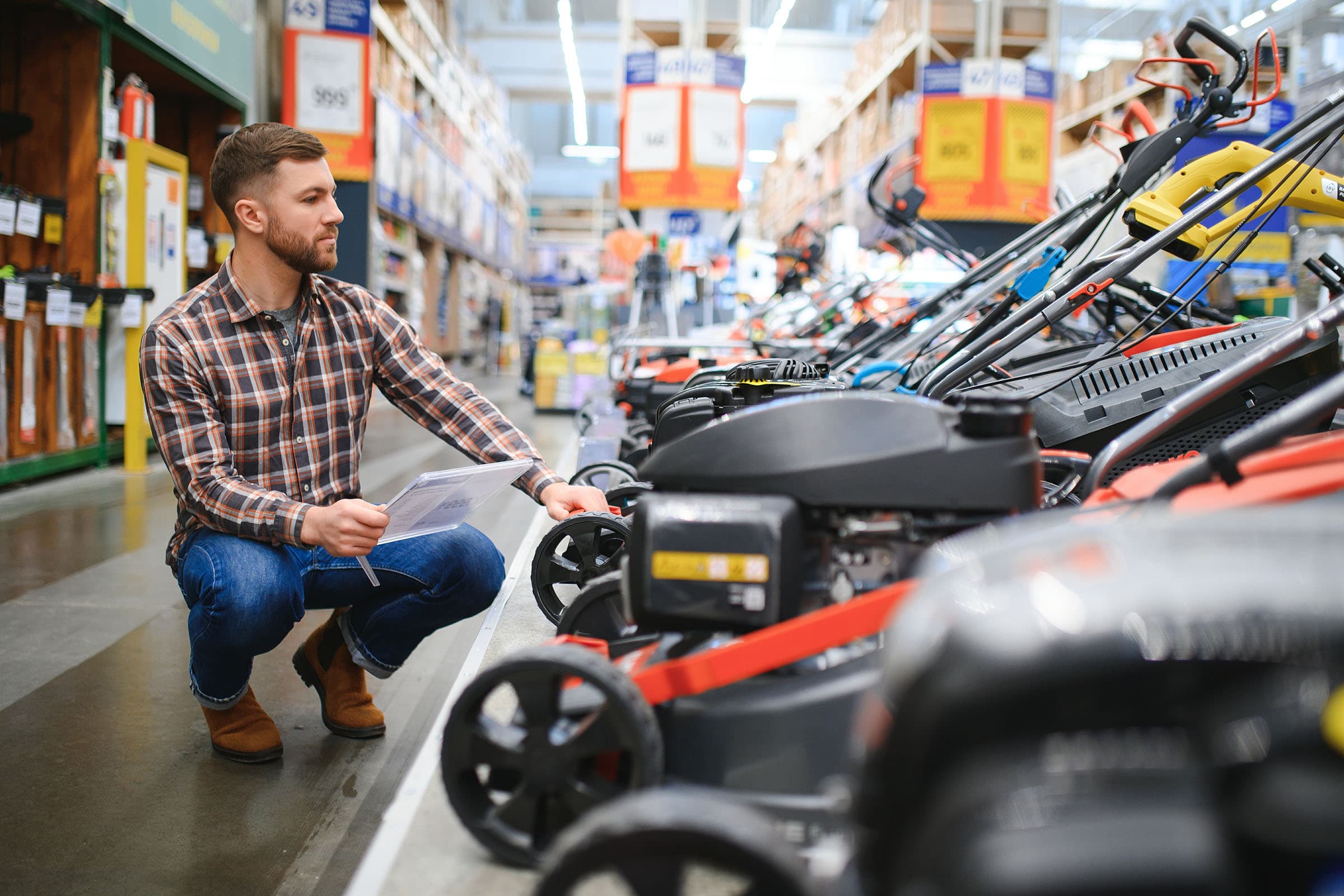 Man in a hardware store kneeling and examining a row of lawn mowers while holding product information sheets.