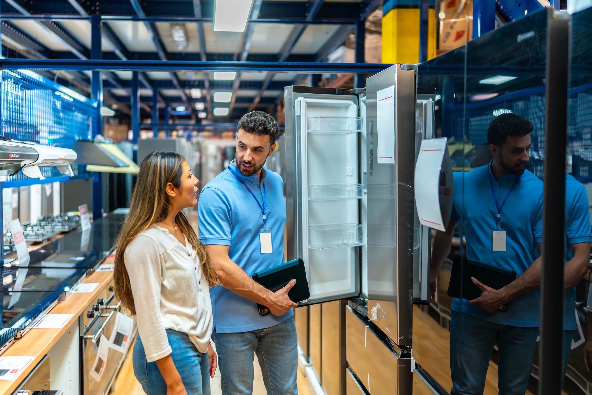 A salesperson shows a customer an open refrigerator in an appliance store, engaging in conversation.