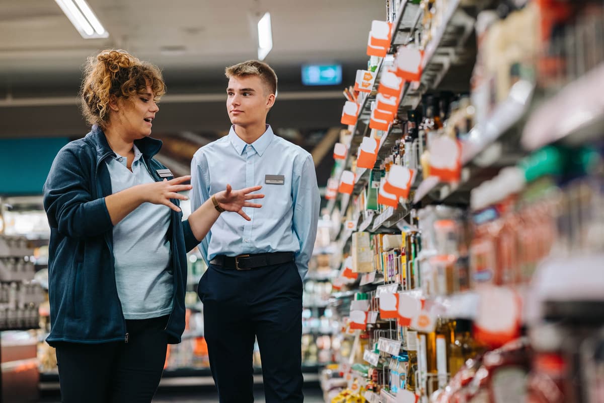 Two store employees in uniform discuss near supermarket shelves, one gesturing with hands while the other listens attentively.