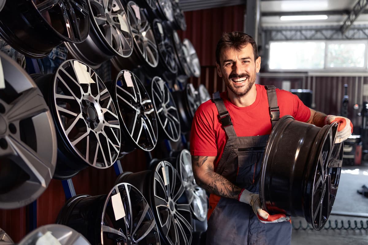 Man in red shirt and overalls holds a car rim, standing in a garage filled with various rims on display.